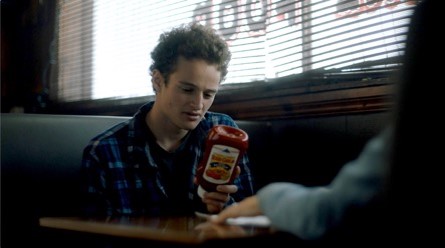 Screenshot from an FOH commercial showing a young man in a restaurant booth looking at a bottle of red gold ketchup